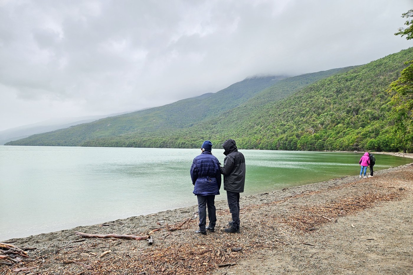 Dos pasajeros de Explore Tierra del Fuego en la costa del Lago Roca en un día nublado, con la nube baja apoyada sobre la ladera del bosque, Parque Nacional Tierra del Fuego
